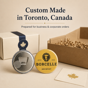 Custom made metal pinback buttons and branded packaging boxes on a beige surface, with text reading 'Custom Made in Toronto, Canada' and 'Prepared for business & corporate orders.' One button is yellow with black BORCELLE BREWERY branding and a maple leaf, and another button shows the silver metal back with a bottle opener cutout.
