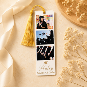 A personalized graduation photo bookmark with a gold tassel, featuring three printed photos of a cap and gown ceremony and the text 'Haley CLASS OF 2026' on a cream background, styled with a cream ribbon, dried flowers, and a wooden tray.