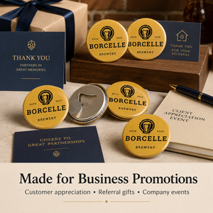 A set of gold and navy blue Borcelle Brewery promotional items, including round button badges, a bottle opener, and matching business cards, arranged on a wooden surface with a gift box and pen.