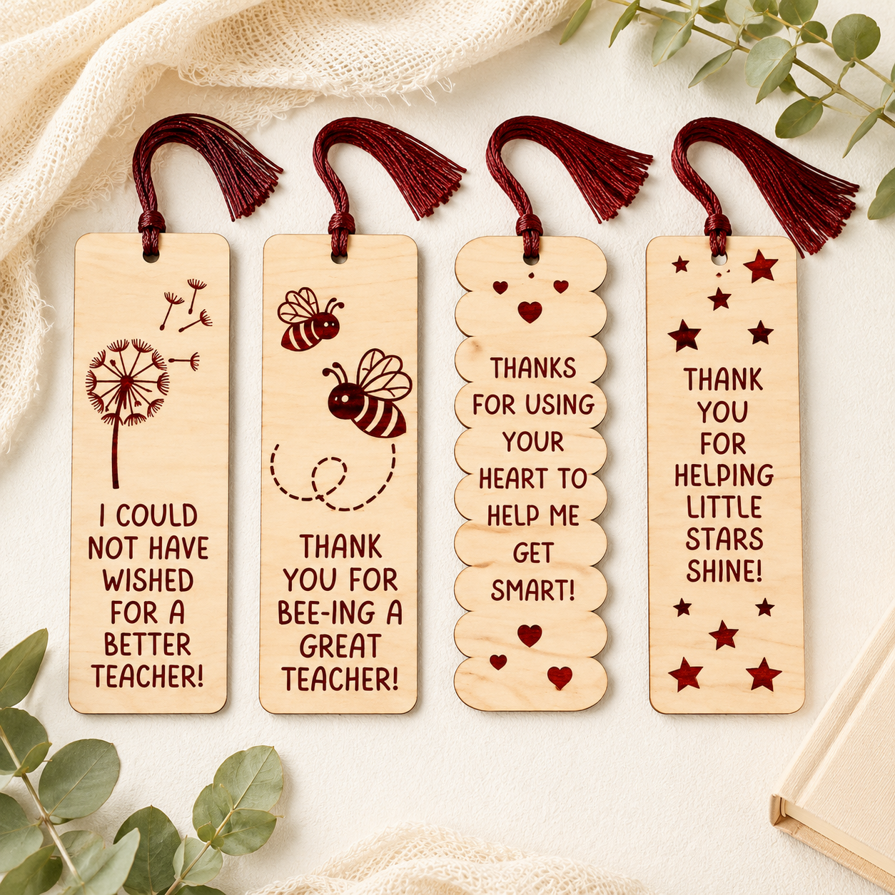 Four personalized wooden bookmarks with dark red tassels, arranged on a cream fabric background with eucalyptus leaves and a book corner. The bookmarks feature engraved designs and messages: a dandelion with 'I COULD NOT HAVE WISHED FOR A BETTER TEACHER!', two bees with 'THANK YOU FOR BEE-ING A GREAT TEACHER!', stacked hearts with 'THANKS FOR USING YOUR HEART TO HELP ME GET SMART!', and stars with 'THANK YOU FOR HELPING LITTLE STARS SHINE!'.